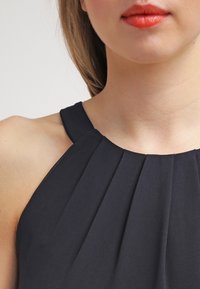 Vestido negro sin mangas con detalles plisados alrededor del escote. La tela parece suave con un ligero brillo, resaltando las curvas.