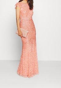 Dlouhé růžové šaty pokryté flitry s krátkými volánkovými rukávy a přiléhavým korzetem, který přechází do rozšířené sukně. Doplněné světle růžovým psaníčkem.