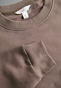 Detailný záber na fialovú mikinu, ktorá zobrazuje rebrovaný kulatý golier, manžety, mäkký fleecový interiér a štítok veľkosti M označený "THE SET", vyrobené na Srí Lanke.