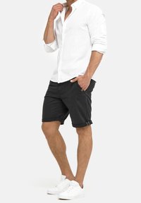 Chemise en lin blanc avec col boutonné, associée à un short noir retroussé et des baskets blanches. Design minimaliste, sans motifs visibles.