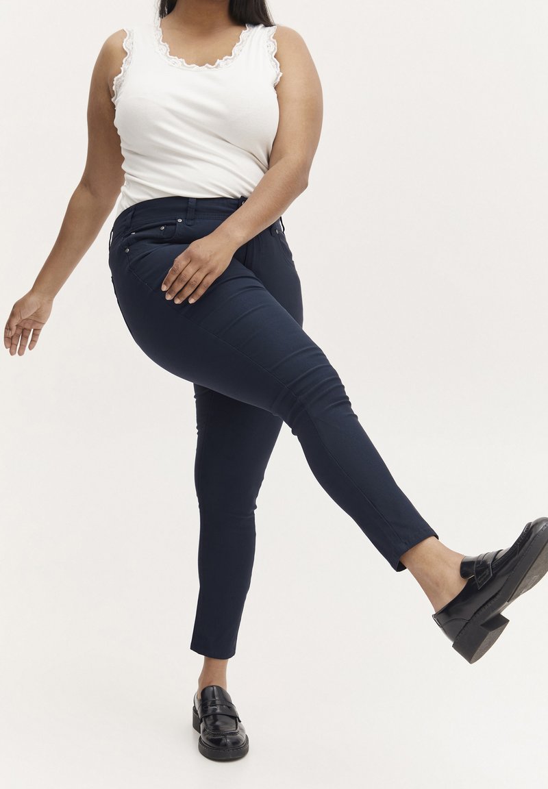 Marineblaue Skinny-Jeans mit glatter Textur, hoher Taille und vorderen Taschen. Kombiniert mit einem weißen Tanktop mit Spitzenrand und schwarzen Slippern.