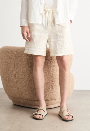 Light beige patterned shorts with an elastic waistband and drawstring, paired with open-toe sandals. Background features a textured, round beige seat.