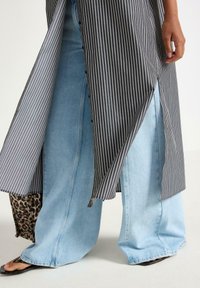 Grå och vit randig skjortklänning lager på lager över ljusblå vida jeans; märkbara leopardmönstrade detaljer vid fållen; platta sandaler synliga.