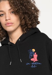 Sweatshirt preto com uma gráfica frontal de uma figura rosa, palmeiras e sol em azul e laranja. Cordões prateados completam o design.