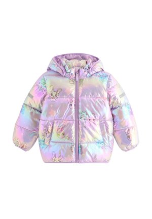 Ung pige i skinnende pastelfarvet jakke med tegneserie-kaniner, blå jeans med kaninprint, lyserød skjorte og lyserøde sko, står og smiler.