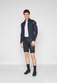 Musta zip-up pyöräilytakki heijastavilla yksityiskohdilla, valkoinen verkkomateriaali sisäpaita, mustat pehmustetut shortsit ja mustat pyöräilykengät tarranauhoilla.