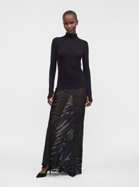 Personne portant un haut noir moulant à col roulé et manches longues avec une jupe longue noire transparente par-dessus un short noir et des talons noirs pointus.