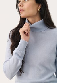 Light blue turtleneck sweater made of knit fabric, featuring ribbed cuffs. The model holds the collar slightly with one hand.