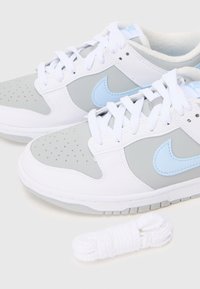 Pár bílých a šedých tenisek Nike s světle modrými logy swoosh a extra bílými tkaničkami na bílém pozadí.