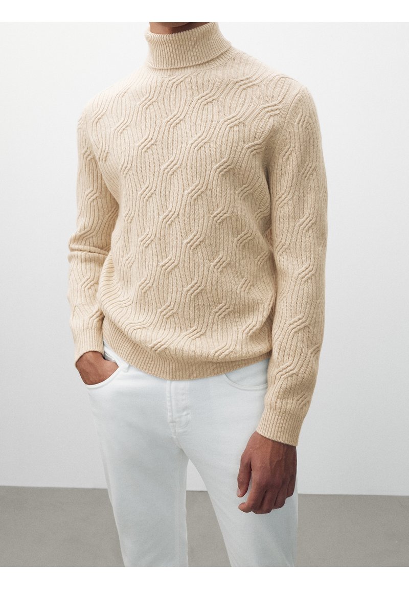 Pull à col roulé beige en maille torsadée porté avec un pantalon blanc, une main dans la poche, fond neutre.