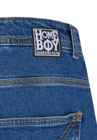 Denim Shorts mit blauer Waschung, gelben Nähten und einem schwarzen Etikett mit weißem Text "HOME BOY loud.couture" am Bund.