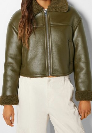 Femme portant une veste en cuir courte vert olive avec col et poignets en peau retournée, associée à un pantalon taille haute écru.