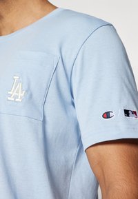 Heledhalli lühikeste varrukatega T-särk, millel on rinnatasku, broderitud LA logo ning väikesed Champion ja MLB logod varrukal.
