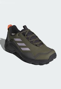 Zapato de senderismo verde oliva con acentos grises, tejido texturizado y suela de goma. Presenta tres rayas moradas y una lengüeta trasera de color naranja brillante.
