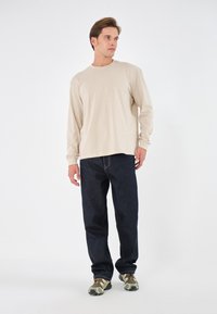 Beige langermet skjorte, mørkeblå jeans og sko med kamuflasjemønster. Antrekket er uformelt med lett stoff og en avslappet passform.