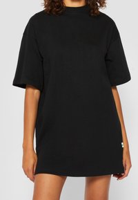 Zwart oversized T-shirt van katoen, met korte mouwen, een hoge halslijn en een klein groen label aan de zijkant. Gladde textuur.