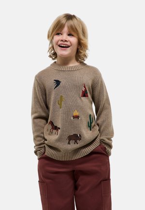 Enfant souriant aux cheveux blonds portant un pull beige avec des motifs western brodés et un pantalon bordeaux, les mains dans les poches.
