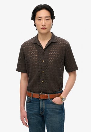 Chemise en tricot à manches courtes, marron foncé, avec un motif en zigzag, dotée d'un col et d'une fermeture à boutons, portée avec un jean bleu et une ceinture marron.