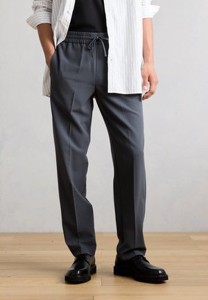 Mann trägt dunkelgraue Hose mit Kordelzug, schwarze Schuhe, schwarzes Hemd und weiß gestreiftes offenes Hemd, steht auf Holzboden.