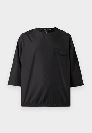 Fekete laza szabású felső könyékig érő ujjakkal, kerek nyakkivágással és egy gombos zsebfedővel a bal mellrészen, fehér háttér előtt.