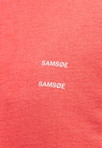 Tissu rouge avec deux logos blancs "SAMSØE" identiques imprimés verticalement, l'un au-dessus de l'autre, centrés dans le cadre.