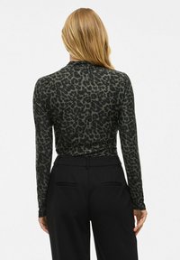 Vrouw met lang blond haar, gekleed in een langwerpige zwarte en donkergroene panterprint top met lange mouwen en hoge zwarte broeken, gezien van achteren.