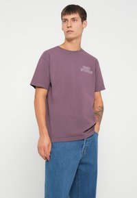 Vijolična bombažna majica s kratkimi rokavi in okroglo ovratnikom, s belim napisom "OBEY STUDIOS" na levem prsnem delu. Nosi se z modrimi jeans hlačami.