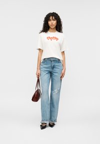 Λευκό κοντομάνικο μπλουζάκι με πορτοκαλί γραφικό "HUGO", συνδυασμένο με ανοιχτό μπλε παντελόνι με φαρδιά μπατζάκια, μαύρα μυτερά παπούτσια και μια μικρή καφέ τσάντα.