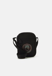 Czarna torba na ramię wykonana z tkaniny, z regulowanym paskiem, z okrągłą łatą z logo w kolorach czerwonym, zielonym i brązowym na przodzie.