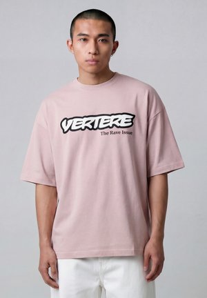 Jonge man met kort haar, gekleed in een lichtroze oversized T-shirt met de tekst "VERTERE The Rave Issue" en een witte broek, tegen een effen achtergrond.