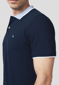 Granatowa koszulka polo z jasnoniebieskim kołnierzykiem i białymi akcentami, z trzema guzikami i teksturowanym materiałem. Subtelne logo na piersi.