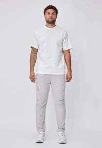 Homme tatoué debout, de face, portant un t-shirt blanc uni, un pantalon cargo gris clair et des baskets blanches, sur un fond uni.