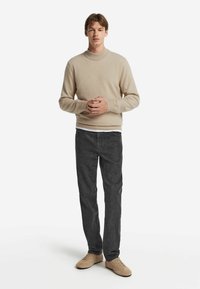 Junger Mann steht, trägt einen beigen Pullover, ein weißes Unterhemd, dunkelgraue Jeans und beigen Freizeitschuhen, mit gefalteten Händen vor der Brust.