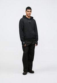 Melns kapuci džemperis ar priekšējām kabatām un aukliņu kapuci, valkāts kopā ar vaļīgām melnām biksēm un pirkstainiem cimdiem. Gluds audums, minimālistisks dizains.