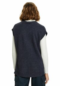 Personne aux cheveux bruns mi-longs portant un gilet sans manches en tricot gris foncé sur une chemise blanche à manches longues, vue de dos.