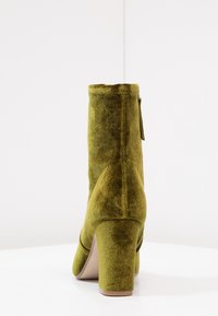Bottine en velours vert olive avec un talon carré, vue de l'arrière sur une surface blanche contre un fond clair.