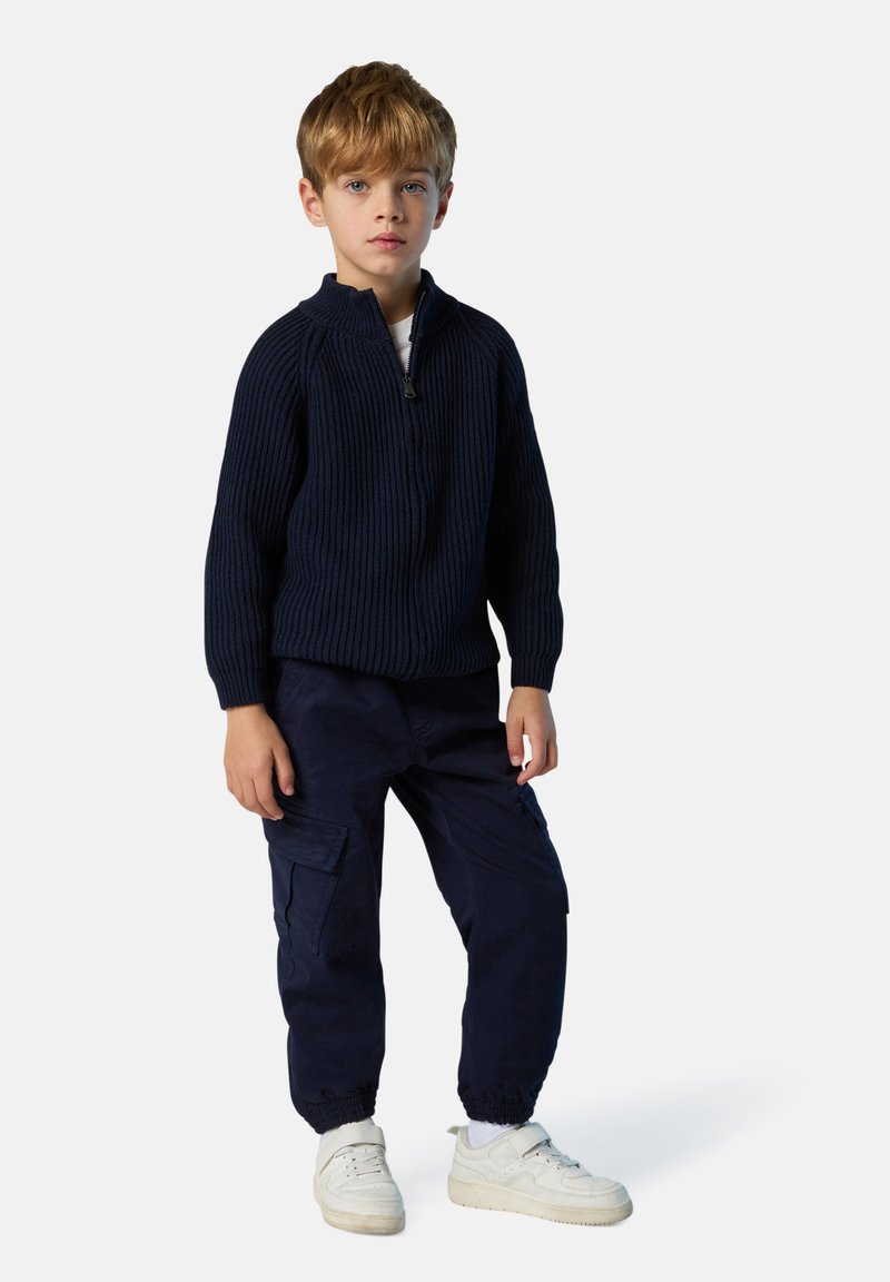 Suéter de punto acanalado azul marino con cuello con cremallera, combinado con pantalones cargo azul marino y zapatillas blancas. Textura suave y corte relajado, diseñado para la comodidad.