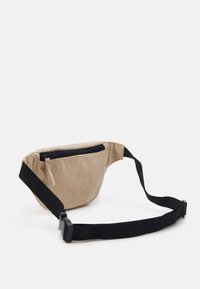 Beige corduroy vyölaukku, jossa on musta vetoketjullinen tasku ja säädettävä hihna; siinä on ribatoitu pinta ja kompakti, pyöristetty muoto.