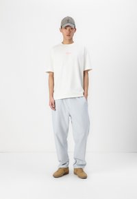 T-shirt beige clair avec un petit logo rose, associé à un pantalon large bleu clair et des baskets beiges. La casquette est grise claire avec une visière courbée.