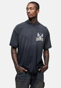 Zwart katoenen T-shirt met een logo met vleugels en tekst, ronde hals, korte mouwen en een relaxed fit; gedragen met donkere spijkerbroek.