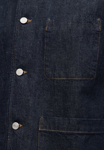 Πουκάμισο από τζιν σε σκούρο μπλε, με ασημένια κουμπιά και πορτοκαλί ραφές. Διαθέτει τσέπη στο στήθος με στρογγυλεμένο κάτω μέρος. Ομαλή υφή.