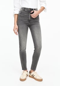 Mujer que viste jeans skinny negros descoloridos de cintura alta, camiseta blanca de manga larga con los puños enrollados y zapatillas color crema con detalles en marrón.