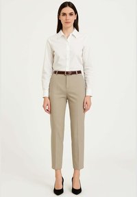 Camicia bianca con bottoni, pantaloni beige su misura, cintura marrone con fibbia argento, scarpe nere con tacco alto, seduta su uno sfondo semplice.