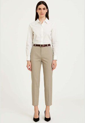 Camicia bianca con bottoni, pantaloni beige su misura, cintura marrone con fibbia argento, scarpe nere con tacco alto, seduta su uno sfondo semplice.