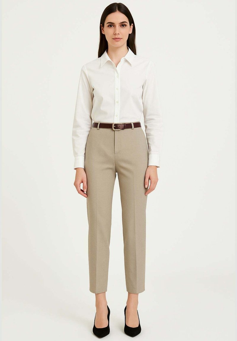 Camicia bianca con bottoni, pantaloni beige su misura, cintura marrone con fibbia argento, scarpe nere con tacco alto, seduta su uno sfondo semplice.