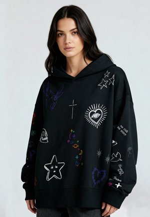 Jonge vrouw die een zwarte oversized hoodie draagt met kleurrijke geborduurde sterren, harten, een oog, een kruis en verschillende abstracte symbolen.