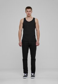 Camiseta sin mangas negra y jeans negros en un modelo masculino, con un diseño ajustado y detalles desgastados en los jeans, combinados con zapatillas negras y blancas.