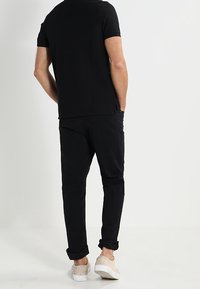 Musta lyhythihainen paita ja mustat housut, esiteltynä takaa. Vaaleat tan-väriset kengät ja käännetyt hihojen päät ovat näkyvissä. Sileä kankaan rakenne.