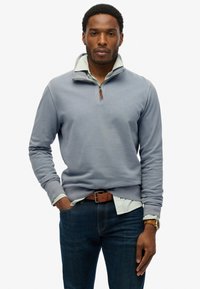 Blauwe pullover trui met kwartrits, geribde kraag en klein logo. Gelaagd over een lichtgroen gestreept shirt, gecombineerd met donkere spijkerbroek.
