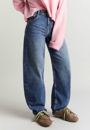 Személy, aki kék, bőszárú farmert, rózsaszín, túlméretezett pulóvert és barna tornacipőt zöld fűzőkkel visel, és egy egyszerű háttér előtt áll.